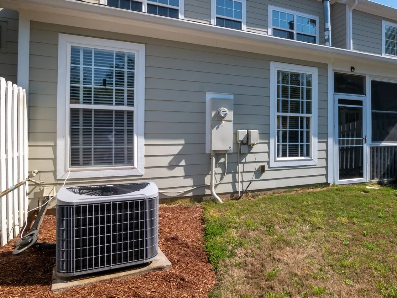 Central air conditioner condenser on a concrete pad in a mulched bed behind a two-story house in summer